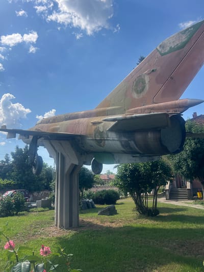 An old, weathered fighter jet is mounted on a concrete pedestal in an outdoor park or public space. The jet, painted in a camouflage pattern of browns and greens, exhibits signs of age and exposure to the elements, with peeling paint and visible wear. Its landing gear is extended, and it appears to be permanently displayed. The surrounding environment includes a well-maintained grassy lawn, several trees, rose bushes in the foreground, and buildings in the background. In the distance, a red car is parked, and there are several large black trash bags on the ground. The sky above is blue with scattered white clouds. This scene is located in the city of Zelenikovo, Bulgaria, where it serves as a monument or public art installation.