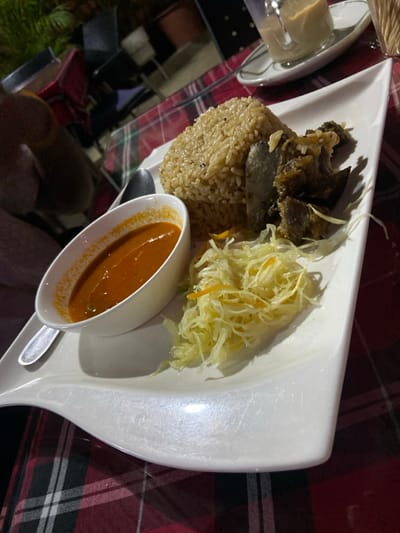 This media file presents a close-up view of a meal served on a modern white plate, set against a red and dark plaid tablecloth. The scene suggests an evening or indoor dining experience in Bagamoyo, Tanzania, given the dim lighting and artificial light reflections.
The main subject is a substantial dish composed of several elements. A mound of light brown, seasoned rice, resembling pilau, occupies the left side of the plate. Resting on and beside the rice are several pieces of dark, cooked meat, possibly stewed beef or goat. To the right of the meat and rice is a portion of finely shredded, pale green or yellow vegetable, likely a cabbage slaw or a simple salad.
A white gravy boat, accompanied by a spoon, holds a rich, orange-red sauce or gravy, possibly a chili sauce or a light curry, and is positioned at the bottom center of the plate. In the upper left corner of the frame, a small glass dish containing a yellowish condiment, perhaps butter, sits on a white saucer.
The image does not show any people, and there is no visible text. The overall impression is that of a rustic yet neatly presented meal, characteristic of local cuisine.