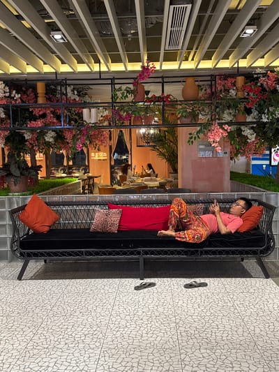 In Nha Trang Ward, Vietnam, a man is lying on a black woven couch, holding a mobile phone. He is wearing a pink t-shirt and colorful patterned pants. The couch is adorned with three pillows: one orange, one with a leopard print, and one red. In the background, a restaurant setting is visible, featuring tables, chairs, and a wall decorated with flowers. The overall environment appears to be an indoor public space with a modern, decorative design.