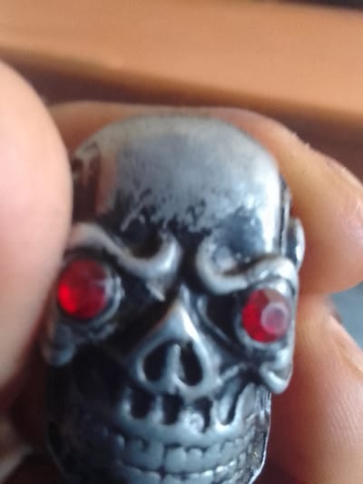 A close-up image, taken in the city of Ndeiya, Kenya, depicts a metallic, skull-shaped object held by human fingers. The object, possibly a ring or charm, is silver-toned with darkened recesses emphasizing its skeletal features. Two round, red gemstones are set into the eye sockets. Portions of two human fingers are visible, with skin texture evident, gripping the object on its left and right sides. The background is out of focus, showing indistinct warm brown and darker tones.