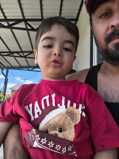 A selfie is taken from a low angle, capturing a man's head and shoulders in the upper right of the frame, and a young child's face in the center. The man is wearing a black tank top and a red baseball cap. The child is wearing a red t-shirt with a graphic of a teddy bear wearing a Santa hat and a scarf. The child's lips are pursed. The background shows a white ceiling with black metal beams, a blue sky with clouds, and a glimpse of houses and trees. The scene appears to be outdoors, possibly on a porch or patio, in the city of Фронтовики Снт, Russia.