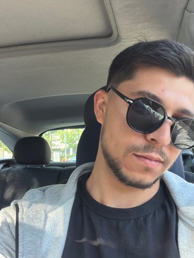A close-up shot looking up at a man in sunglasses. He is seated in a car, visible by the interior details such as the headrest and sun visor. Outside the car window, trees can be seen, indicating a location with greenery. The man has dark hair and a beard. He wears a black t-shirt and a gray hoodie. The city of Gütersloh, Germany, cannot be confirmed from the image.