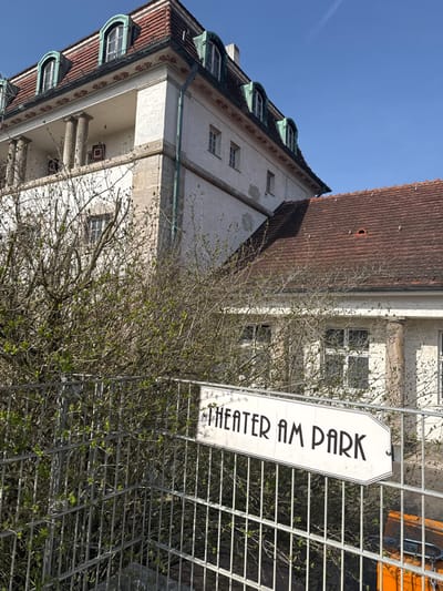 The image displays an exterior view of a building identified as "Theater am Park" in Bad Nauheim, Germany, under a clear blue sky. A white directional sign with a black border and "THEATER AM PARK" written in black, Art Deco-style font is affixed to a galvanized metal fence in the foreground, pointing right. Behind the fence, deciduous shrubs with nascent green leaves partially obscure the lower facade of the building. The structure features multi-story white textured walls and a prominent red-tiled roof. The upper section of the building includes a balustraded balcony supported by stone columns and multiple dormer windows with distinctive arched, green-patinated copper frames. Rectangular windows are visible on the lower section of the building. A partially obscured orange object is visible behind the fence in the lower-right corner. No individuals are present in the scene.