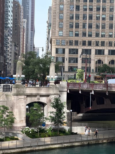 The image depicts a view of the Chicago River in Chicago, Illinois, USA. In the foreground, the river is visible with its greenish-blue water. A concrete embankment lines the river, topped by a metal railing. Beyond the railing, a paved walkway curves gently. On this walkway, three individuals are present: two women appear to be conversing, and a woman wearing a white hat stands facing away from the camera.
Behind the walkway, a section of a stone bridge pier is visible, featuring arched openings and decorative stonework. Further back, a tree-lined area with white flowers adds a touch of greenery. Several trees stand on a lower level, casting shadows.
Above the bridge pier, a multi-story building with a light-colored stone facade and numerous windows dominates the midground. A traffic light with red and yellow signals is visible on the left side of the frame. A street sign indicating "Wabash Ave" is also present. A dark red bridge with a decorative metal railing spans across the river in the midground.
In the background, a dense urban landscape of tall buildings with various architectural styles and materials rises into a hazy sky. Some buildings have a grid-like pattern of windows, while others feature decorative elements. The overall scene suggests a sunny day in a bustling urban environment.