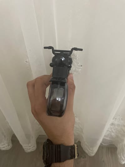 A light-skinned human hand, exhibiting forearm hair, holds a black, motorcycle-shaped spray bottle vertically against a light-colored, sheer fabric background. The hand grasps the bottle with fingers wrapped around the transparent dark gray lower section, which contains a clear liquid, and the thumb partially visible near the top opaque black section. A black digital watch with "PROTECTION" visible on the strap and a gold-colored buckle is worn on the wrist. The bottle is designed with distinct handlebars and engine block details. The background fabric, likely a curtain or drape, features a fine mesh texture and appears to have embroidered or lace detailing, particularly visible on the right and lower sections, with vertical folds and soft shadows. The image contains no discernible features to indicate a specific geographic location, including Ihtiman, Bulgaria.