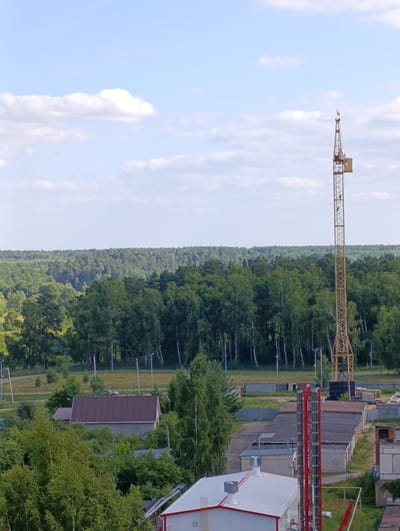 This is a medium-angle, high-shot photograph taken on a bright, overcast day. The scene is a construction or industrial area situated at the edge of a dense forest. Dominating the right side of the frame is a tall, yellow lattice tower crane, typical for construction sites. Below and to the left of the crane, various industrial buildings and structures are visible. These include a long, low-rise building with a gray roof and another with a darker roof. In the foreground, there's a modern-looking building with a white roof and red trim, and prominent red vertical pipes, possibly part of an industrial ventilation or exhaust system. A smaller, older building with a brown, pitched roof is nestled among trees to the left.
The background is filled with a vast expanse of lush green forest, characteristic of temperate climates. The sky above is a pale blue with scattered white, fluffy clouds, suggesting it is daytime, likely mid-morning or afternoon, with mild weather. No people are clearly visible, but the presence of the crane and ongoing construction implies activity is taking place. The overall impression is one of a functional, industrial zone operating near a natural environment. The location is identified as Alexandrov, Russia, which aligns with the typical architecture and landscape. No discernible text is present in the image.