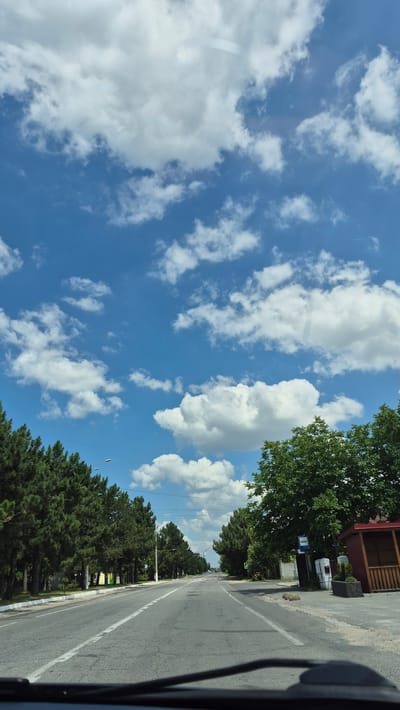 The image shows a straight, paved road lined with trees on both sides. The road has white lane markings. On the right side of the road, there is a bus stop shelter and a blue bus stop sign. The sky is blue with scattered white clouds. The overall scene appears to be a quiet street in a suburban or semi-rural area. Based on the available visual information, the location is identifiable as a street in the city of Ialoveni, Moldova.