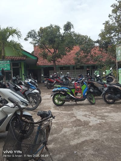 A parking area is filled with motorcycles. A building with a red tile roof and green trim stands behind the vehicles. Trees are interspersed throughout the scene. A bicycle with a basket in the foreground is partially obscuring the view. The pavement appears to be cracked and worn. The sky is overcast. A timestamp indicates November 11, 2025.