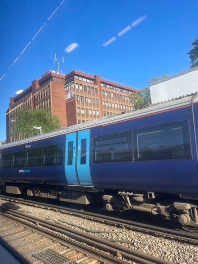 The image, originally oriented 90 degrees clockwise, captures a bright, sunny daytime scene from the perspective of a train window. The clear blue sky dominates the upper and left portions of the frame, showing faint reflections of interior lighting on the glass.
On the right side, a dark blue Southeastern train is prominently featured, identifiable by its "southeastern" branding and unit number "74910". It has contrasting light blue doors. Below the train, detailed railway tracks with gravel ballast are visible, along with some of the train's mechanical undercarriage components.
In the background, a tall, multi-story building constructed of reddish-brown brick stands against the sky on the left. It features many windows and what appears to be scaffolding or an antenna array near its roofline. A large banner on the building advertises "BRAND NEW LUXURY STUDIOS AND ONE BED APARTMENTS AVAILABLE TO RENT" and provides contact numbers: "020 3230 0080" and "07711 590 719". Green foliage from trees is visible between the train and the building. The scene conveys a clear, bright day in Tonbridge and Malling, United Kingdom.