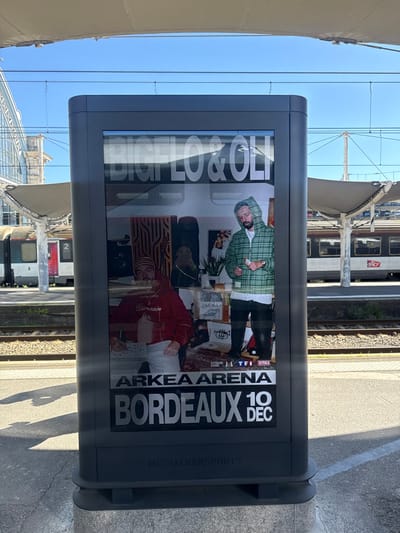 The image depicts a vertical, grey advertising display positioned on a concrete train station platform under clear blue skies. The display features a poster for "BIGFLO & OLI" performing at "ARKEA ARENA BORDEAUX 10 DEC." The poster shows two men; one on the left wears a red hoodie with "Billionnaire" text and a striped beanie, looking down. The other man on the right wears a green plaid hoodie and black pants, also looking down at an object in his hands.

In the background, train tracks run horizontally, separating the platform with the display from other platforms and railway infrastructure. To the left, a white and red train car (partially visible, likely SNCF) is parked at an adjacent platform, with an ornate, light-colored building (characteristic of 19th-century train station architecture) visible beyond it. To the right, another white and red train (with "SNCF" visible on its side in the crops) is parked on a parallel track. Both platforms are covered by light-colored canopy structures, supported by poles. Overhead catenary wires are visible against the bright blue sky. Long shadows are cast on the concrete platform, indicating direct sunlight from a high angle.

This scene is located within a train station in Bordeaux, France, as indicated by the poster promoting an event in "Bordeaux" and the presence of SNCF trains.