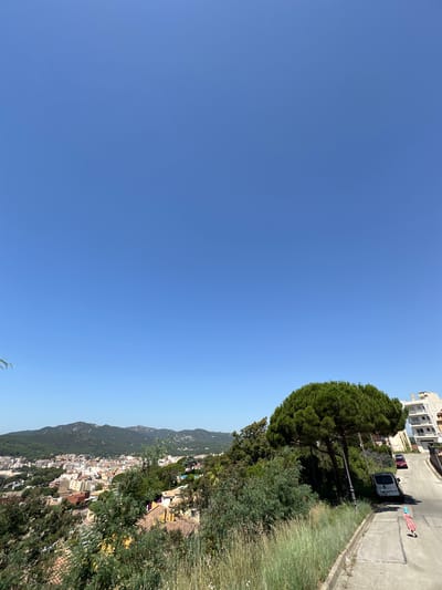 The image captures a sun-drenched day in Sant Feliu de Guíxols, Spain, with a clear, expansive blue sky dominating the upper portion of the frame. In the foreground, a paved pathway slopes downwards, bordered by tall, dry grasses and a large, dense pine tree with a rounded canopy. To the right of the pathway, a white building with multiple balconies is visible. On the pathway itself, a silver van is parked, and further down, a person wearing a red dress and a striped scarf is walking away from the viewer. Two cars, one red and one dark-colored, are parked on the street beyond the pathway.
In the middle ground, a panoramic view unfolds of the coastal town of Sant Feliu de Guíxols. Buildings with terracotta roofs and white facades are clustered together, nestled against a backdrop of lush green hills covered in trees. The distant mountains form a rugged horizon against the clear sky. The overall impression is one of a bright, summer day in a Mediterranean coastal town.