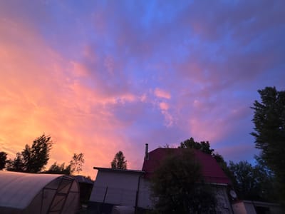 The image depicts a rural scene in Zaprudnya, Russia, at dusk. The sky is filled with dramatic clouds displaying a gradient of colors, transitioning from fiery orange and pink on the left to deep blue and purple on the right. In the foreground, a white greenhouse with a metal frame is partially visible on the left. To the right of the greenhouse, a two-story brick building with a red tiled roof stands prominently, featuring a chimney and a satellite dish on its side. Silhouettes of trees are scattered throughout the scene, adding depth and context to the rural environment. The lighting suggests the sun has recently set, casting a warm glow on the clouds and illuminating the scene.
