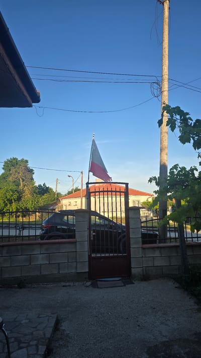The image captures a scene in Rudnik, Bulgaria, on a clear day. A Polish flag, torn and weathered, flies from a flagpole in front of a house with a red-tiled roof. The house is behind a metal gate and a low brick wall. A black car is parked behind the gate. The foreground shows a stone-tiled pathway and a gravel area. Power lines stretch across the sky, and a concrete utility pole stands to the right, partially obscured by leafy branches. The overall impression is one of a residential area in daylight.