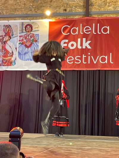 The image shows a performance at the Calella Folk Festival in Calella, Spain. A person dressed in traditional dark clothing with red accents is captured in mid-leap on a stage. They have long, dark hair that obscures their face, and are wearing boots and a belt with a buckle. The backdrop features a large red banner with white text that reads "Calella Folk Festival". Below the banner, there are posters depicting people in colorful traditional costumes. The stage is set against a dark curtain, and to the right, a wooden wall is visible. A bright light is visible in the upper left corner, and a camera is in the lower right corner. The scene suggests an energetic and celebratory folk music or dance performance taking place indoors. The overall impression is one of dynamic movement and cultural display.