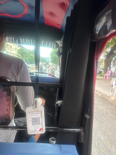 The image shows the interior of a tuk-tuk, a three-wheeled motor vehicle commonly used as a taxi. The tuk-tuk appears to be in motion on a city street, suggested by the blurred background and the presence of other vehicles and pedestrians. The interior is upholstered in red and blue. Hanging from a lanyard is a white card with a QR code and the text "ABA PAY Scan For Done". Below the QR code, a name is visible, "THORN YA OU", and a merchant ID, "MID: 12309291045091". The card also displays the KHQR logo and contact information. A person, likely the driver or a passenger, is visible on the left, wearing a white shirt. The environment outside the tuk-tuk suggests a warm climate with green trees and buildings. The location is identified as Khan Prampir Makara, Cambodia.