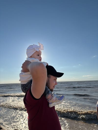 In Port-Katon, Russia, a man wearing a maroon t-shirt and a black baseball cap carries a baby on his shoulders. The baby, adorned with a white turban, gazes towards the ocean. The sun is visible behind the baby, casting a bright glow. The water's surface reflects the sunlight, creating a shimmering effect. The scene suggests a family enjoying a day at the beach.
