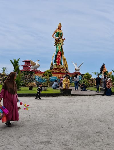 The image depicts a sunny day at a public square in Kota Pari, Indonesia, featuring a large, vibrant statue of a woman in traditional attire. The statue is adorned with green and red fabric, with gold accents, and is positioned on a raised platform. Flanking the central statue are two large, white swan statues with yellow beaks and wings, one on either side. In the background, a red and white tiered building with white ornamental birds is visible. A group of people are gathered around the statues, some seated, some standing, and some taking pictures. In the foreground, a woman in a pink hijab and a long pink dress is walking from left to right, carrying a collection of colorful pinwheels and other toys. A young boy in a black shirt and pants is running in the middle ground. The ground is a light-colored gravel path. The sky is partly cloudy with a clear blue expanse.