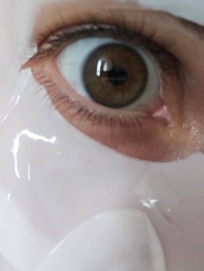 A close-up, macro photograph captures a human eye, likely of a female, with a brown iris. The eye is partially covered by a white, translucent material, possibly a facial mask or sheet. The skin around the eye appears smooth, with visible eyelashes. The background is out of focus and dominated by the white material, reflecting light in several areas, suggesting a shiny or wet surface. The lighting is soft and diffused. The scene offers no contextual clues about its location in Petropavlivska Borshchahivka, Ukraine, or any other specific environment.