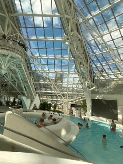 This photograph shows a water park located in Les Escaldes, Andorra. The park features a large, glass-domed roof with a white, geometric support structure. The sky is visible through the glass, showing scattered clouds. Inside, there is a pool with several people swimming and standing in the water. A white water slide is partially visible on the right side of the image. There is a large black screen mounted on the wall. Some green plants are visible near the pool area. The image is taken from an angle looking upwards towards the roof and downwards into the pool area.