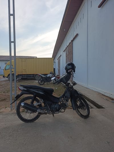 A black motorcycle is parked on a dirt and concrete surface in front of a large, white warehouse. A black helmet with a "HONDA" logo rests on the handlebars of the motorcycle. In the background, a yellow truck is parked next to another motorcycle. The warehouse has a corrugated metal roof and large sliding doors. The sky is cloudy. The scene is located in the city of Dumai, Indonesia.