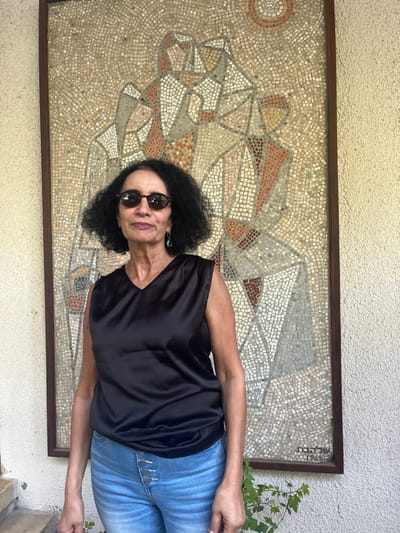 A single adult female subject is depicted standing and facing the camera outdoors in the Brener Regional Council, Israel. She appears to be in her 50s or 60s, with dark, curly hair and medium skin tone. She wears dark, round-framed sunglasses, a black V-neck sleeveless top, and light blue jeans featuring four prominent buttons on the fly. A light blue, teardrop-shaped earring is visible on her left ear. Behind the subject is a large, rectangular mosaic artwork mounted on a light-colored, textured wall. The mosaic, framed by a dark brown border, comprises small tesserae in shades of beige, white, light grey, brown, and reddish-orange, depicting abstract, stylized human-like figures and a circular motif at the top. The Hebrew word "שלחבת" and the year "1963" are visible in the bottom right corner of the mosaic. A small green plant is partially visible in the bottom right corner of the frame. The scene is brightly illuminated by natural light, suggesting an outdoor or open-air setting.
