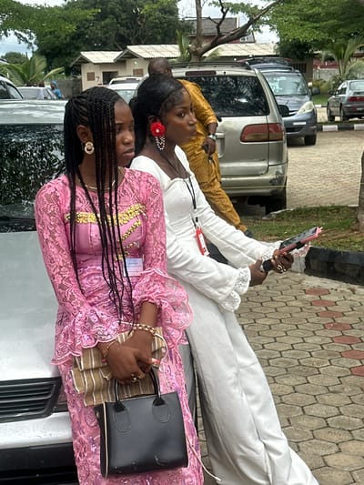 In Itoga, Nigeria, two young women are posed by the side of a silver car. The woman on the left, wearing a pink lace dress with gold embellishments, has her hair in braided extensions and is holding a black handbag. The woman on the right wears a white dress and is holding a mobile phone with a pink case, and a microphone. They appear to be at an outdoor event, with other vehicles and buildings visible in the background. The ground is paved with hexagonal tiles.
