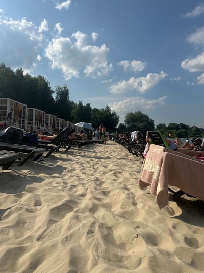 The image shows a sandy beach with several people relaxing on lounge chairs. The scene is set on a sunny day, with a bright blue sky dotted with white clouds. Trees line the background, suggesting a natural or park-like setting. Many of the people are dressed in casual beachwear, and some are facing the water, while others are engaged in conversation. A pink towel is draped over a lounge chair in the foreground, adding a splash of color to the sandy landscape. The sand itself is textured with ripples and shadows, indicating the play of sunlight. The overall mood appears to be one of leisure and relaxation. While the exact time of day is not specified, the strong sunlight and shadows suggest it is likely afternoon. The location is identified as Dal'nie Prudishchi, Russia, which aligns with the visual cues of a beach and surrounding trees. There is no visible text in the image.