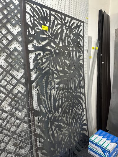 The image depicts an indoor scene, likely a retail or storage space, in the city of Dub, Montenegro. The primary focus is on several large, decorative metal screens with intricate cut-out designs, primarily featuring leaf motifs.

To the left, a metal screen with a geometric diamond pattern is visible. Adjacent to it, a larger metal screen with a more organic, leaf-like pattern dominates the center. This screen has a yellow price tag affixed to it, which reads "135,00 €". Behind these screens, a white pegboard with small, evenly spaced holes serves as a backdrop.

Further to the right, a stack of dark-colored metal sheets or panels stands vertically. In front of these panels, two narrower, lighter-colored perforated metal sheets are leaning. The lower portion of the image shows a stack of blue and white boxes of welding electrodes. The branding "EZ" is visible on the boxes, along with text in a foreign language that appears to be "WELDING ELECTRODES".

The lighting is even, suggesting an indoor environment with artificial lighting. The overall impression is one of a display or inventory of metal products, possibly for sale or in storage. There are no people visible in the image. The environment appears to be a commercial space, possibly a hardware store or a fabrication workshop.