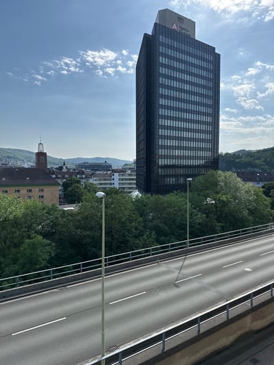 The image depicts a view of Hagen, Germany, from an elevated position, possibly a bridge or high building. A multi-story office building, identified by the logo "Agentur für Arbeit" (Employment Agency), dominates the left side of the frame. The building is of modern design with a grid pattern of windows. To the right of the building is a roadway with multiple lanes and road markings. Between the building and the road is a line of trees and the streetlights. In the background, there are buildings of varying heights and a hilly, wooded area. The sky is blue with scattered clouds. The light suggests it's daytime.
