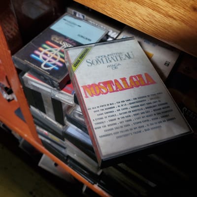 A close-up shot shows a collection of audio cassette tapes stored in what appears to be a cabinet or shelf. Prominently displayed is a cassette titled "Nostalgia" by "Orquestra Som Bateau Ataca de", with the song list partially visible on the cover. The other tapes include "Serie Medium" and an "Classics" tape. The environment suggests an indoor setting. The specific location in Curitiba, Brazil cannot be determined from this single image.
