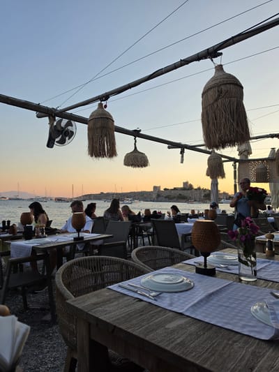 This media captures an outdoor dining scene in Bodrum, Turkey, during what appears to be sunset or twilight. The image is oriented vertically, with the sky and a body of water in the background, dotted with numerous sailboats. The foreground features several tables set for dining, with checkered tablecloths, plates, and cutlery. Wicker chairs surround the tables, and some patrons are visible seated and engaged in conversation or dining.
Above the tables, decorative elements made of straw hang from poles, creating a rustic and natural ambiance. A large fan is also attached to one of the poles. The lighting suggests it's late afternoon or early evening, with a warm, golden glow from the setting sun illuminating the scene. The presence of the sea and sailboats strongly suggests a coastal or harbor-side location. The overall atmosphere seems relaxed and pleasant, with people enjoying an evening meal outdoors. No visible text is present in the media.