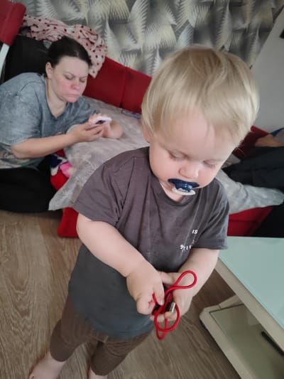 A light-skinned toddler with blonde hair and blue eyes is shown from the chest down, looking down at a red cord in their hands. The toddler wears a dark gray t-shirt and brown leggings, with a blue pacifier in their mouth. The background features a woman with dark hair, wearing a gray t-shirt, looking at a pink smartphone while lying on a red couch. The scene is indoors, with a wooden floor and a wall with a patterned wallpaper. The image was taken in Gvardeysk, Russia.