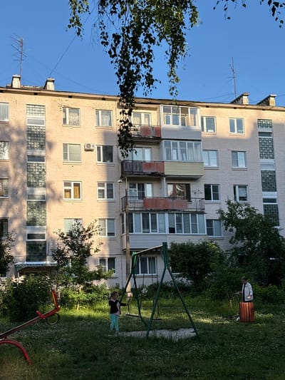 The image shows a side view of a multi-story apartment building, likely in Sarov, Russia, under a clear blue sky. The building is made of light-colored brick and has many windows and balconies. Some balconies are enclosed with glass, while others appear to be open. On the roof of the building, there is a television antenna.
In front of the building, a grassy area with trees and a playground can be seen. A child is visible on a swing set, appearing to be playing. A red seesaw-like toy is also present in the foreground. The sunlight suggests it is daytime, possibly late afternoon, as indicated by the shadows cast by the trees on the building. The overall scene depicts a residential urban environment with a playground, suggesting a typical neighborhood setting. There is no visible text in the image.