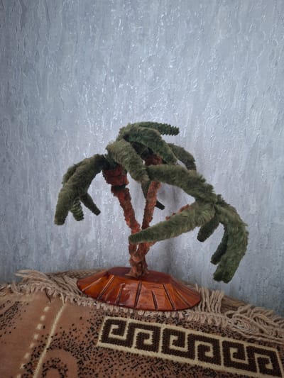 The image shows a close-up, upward-facing shot of a decorative object against a textured, pale blue background. The object appears to be a faux plant with fuzzy, green, branch-like appendages that radiate outwards. Some of these appendages have a rougher, darker green texture at their tips, resembling small cones. The central structure of the plant is wrapped in a reddish-orange fuzzy material, suggesting a twisted stem or vine.

To the right of the plant, a portion of a woven tapestry or rug is visible. This textile is primarily a light brown color with a repeating geometric pattern in dark brown, resembling a Greek key motif. The edges of the tapestry are fringed with light brown yarn. Partially obscured by the plant and tapestry is a carved wooden element, possibly part of a decorative stand or base. It is orange-brown in color and has a fan-like shape with incised lines.

The background has a mottled, uneven texture with lighter and darker patches, possibly representing a wall or ceiling with an interesting finish. The lighting appears to be diffused, casting no harsh shadows. The scene is static, with no obvious activity taking place. The image does not contain any visible text. The orientation suggests the image was taken at an angle, possibly looking up at the object. The overall impression is of an indoor decorative arrangement, possibly with a rustic or folk art aesthetic, given the woven tapestry and carved wood.