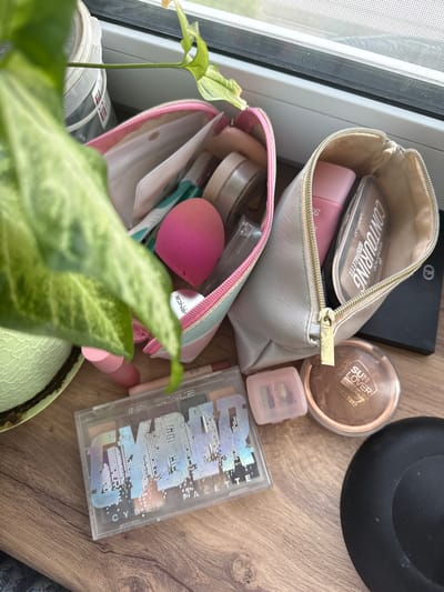 The image shows a collection of makeup products and a potted plant on a wooden surface, likely a windowsill. The makeup items include a pink makeup sponge, a contouring palette, bronzer, lipstick, and an eyeshadow palette. The eyeshadow palette is clear with colorful shades and a cityscape design. The plant appears to be a houseplant with large green leaves. The background suggests an indoor setting with a window. The scene is lit by natural light.