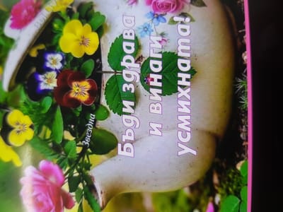 The image displays a close-up of a decorative ceramic pot filled with various colorful flowers, predominantly pansies and roses. The pot itself is light-colored, possibly white or cream, with a slightly glossy finish. It appears to be a decorative item, perhaps a planter or a vase, with some subtle text printed on its surface. The flowers are vibrant, showcasing shades of yellow, purple, red, and pink. Green leaves and stems intertwine with the blossoms, creating a lush and full arrangement. In the background, the environment is blurred but suggests an outdoor setting, possibly a garden or a balcony, with hints of greenery and possibly other floral elements.

The text on the pot is in Cyrillic script. The prominent text reads "Бъди здрава и винаги усмихната!" which translates to "Be healthy and always smiling!". Another smaller text, "Звезднa", meaning "Star", is visible on the side of the pot, along with what appears to be a small ladybug on a leaf.

The overall impression is one of a cheerful and aesthetically pleasing composition, likely intended as a greeting card or a decorative piece. No specific location within the city of Lomets, Bulgaria is identifiable from the image itself.