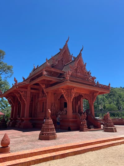 An ornate, bright red Buddhist temple stands under a clear blue sky in Baan Hua Thanon, Thailand. The temple features intricate carvings and decorative rooflines with tiered spires. In front of the temple, a paved courtyard is decorated with red brick patterns and several ornamental red statues. A person in a white shirt and blue shorts stands on the steps to the temple, and another person in a white shirt is visible further back. To the left of the temple, a lush green tree adds a natural element to the scene, and a hill covered in dense vegetation rises behind the temple on the right. The overall impression is one of vibrant color and detailed craftsmanship in a warm, sunny environment.