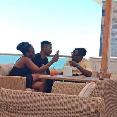 Three individuals, two males and one female, are seated at a table outdoors, with the sea and a clear blue sky in the background. The woman, on the left, has distinctive teal-colored braids piled high and is wearing a black tank top and sunglasses. She is holding a smartphone, seemingly showing it to the man next to her. The man in the middle, wearing a black t-shirt, is looking at the phone and holding a drink with a straw. The man on the right, wearing a pale yellow t-shirt and glasses, is looking at the man in the middle and holding a drink. Two colorful drinks with straws are visible on the table, along with a white sugar dispenser and other items. The seating appears to be wicker furniture, suggesting a casual outdoor dining or lounge area. The location is stated to be in Shchyolkine, Ukraine.