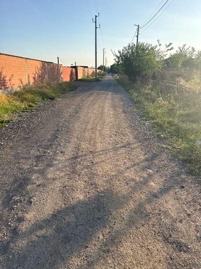 The image depicts a dirt road in Shishmanți, Bulgaria, with a clear blue sky overhead. On the left side of the road is a long, orange brick wall. On the right side, there are overgrown weeds and trees. Several utility poles with power lines are visible along the road. The road itself is unpaved and appears to be a residential access road, with tire tracks visible on its surface. Shadows from the trees cast dappled patterns on the road, indicating it is late afternoon or early morning. In the distance, a person is walking away from the camera on the road. The overall scene is a rural or semi-rural setting.