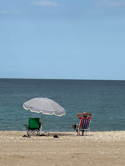 The image depicts a beach scene in M'Diq, Morocco. A man sits in a striped beach chair at the water's edge, hands behind his head. Next to him, on the beach, is a striped beach umbrella and a green folding chair. The scene is split between the clear blue sky to the left and the darker water and beach area to the right. The beach is made of sand and small shells.