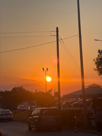 A telephoto shot captures a sunset scene in Keramoti, Greece. The sun, a bright orb, is positioned in the upper center-left of the frame, casting a warm orange glow across the sky and clouds. Two utility poles stand prominently in the foreground, with power lines crisscrossing between them. In the middle ground, trees and vegetation line a street where several cars are parked. A street lamp with two lights is visible near the sun. In the background, there is a glimpse of what appears to be a harbor or marina with boats, and a white tent-like structure. The overall impression is a coastal town bathed in the soft light of dusk.