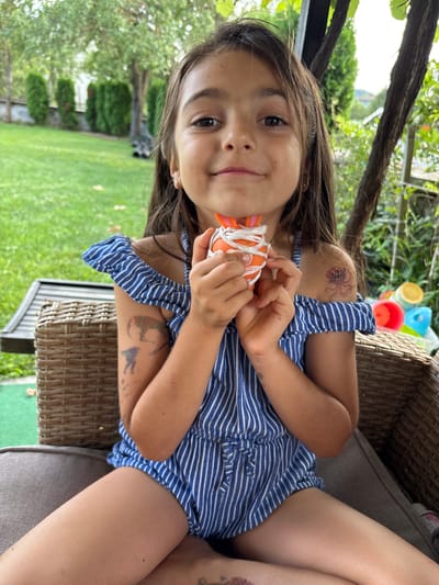 In Oreshak, Bulgaria, a young girl with long brown hair is seated on a wicker chair outdoors. She is wearing a blue and white striped romper. Her left arm is extended towards the camera, holding a small orange object wrapped in white string. She has temporary tattoos on her arms, including a red octopus on her left shoulder and what appears to be a silhouette of an animal on her right arm. She is smiling and looking at the camera. In the background, lush green trees and a lawn are visible, suggesting a sunny day. To the right of the girl, colorful plastic cups are stacked, possibly toys or part of a game. The overall scene conveys a sense of playful outdoor activity.