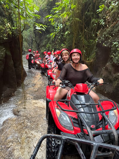 A group of approximately nine individuals wearing red helmets are riding red All-Terrain Vehicles (ATVs) through a shallow, flowing river in a narrow, lush jungle canyon located in Payangan, Indonesia. The immediate foreground features the front of a red KYMCO ATV, with a woman in a black loose-knit top and shorts driving, smiling towards the camera. A second woman, also smiling, is seated behind her as a passenger. Behind them, a line of other red ATVs proceeds through the river, occupied by additional riders and passengers. One individual further back, near the left canyon wall, is standing in the river next to an ATV, wearing black clothing and a black backpack, possibly a guide. The river water is turbulent and light brown, reflecting some ambient light, with splashes around the ATV wheels. Both sides of the narrow passage are defined by steep, dark rock walls heavily covered in dense green tropical foliage, moss, and numerous long, thin hanging vines descending from above. The overall scene depicts an organized recreational activity within a natural, enclosed environment.