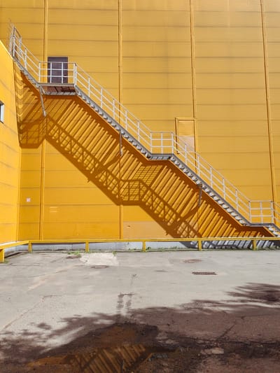 The image shows an exterior view of a bright yellow, corrugated metal building. A metal fire escape with white railings ascends diagonally across the face of the building. The fire escape casts a strong, linear shadow on the yellow wall. At the top of the fire escape, a dark-colored door with a small window and a red handle is visible. Below the fire escape, a yellow metal railing runs along the base of the building. The ground in front of the building is a cracked concrete surface with a small puddle reflecting the shadow of the fire escape and some nearby foliage. The setting appears to be an industrial or commercial area, likely within the city of Putilkovo, Russia, given the context. There are no people or animals present in the image.