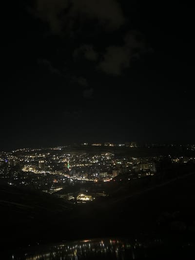 The image displays a panoramic nighttime view of a city, identified as Dummar Municipality, Syria, observed from an elevated vantage point. The upper portion of the frame shows a dark sky containing indistinct, darker grey cloud formations and faint star points. The middle ground is dominated by the sprawling city, illuminated by numerous artificial lights from buildings and streets, appearing as dense clusters of bright white, yellow, and occasional red and green points. A distinctive green-lit minaret is visible within the central-left area of the urban expanse. The city lights extend across undulating terrain, suggesting a valley or hillsides. The foreground consists of dark, unlit landforms (likely hills or slopes) framing the lower part of the cityscape. At the very bottom of the image, a body of water is present, reflecting the city lights as blurred, shimmering patterns on its dark surface. No individuals or specific actions are discernible. The environment is urban at night, with partial cloud cover.