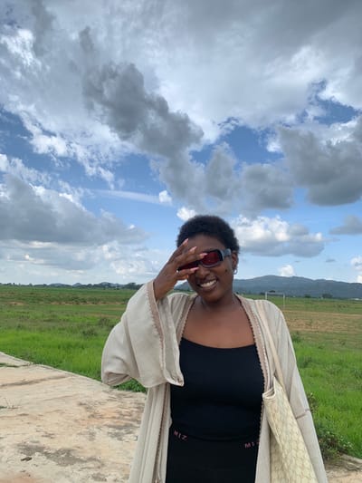 A young Black woman is pictured outdoors, smiling and looking towards the camera. She is wearing a black tank top and a light-colored open cardigan with a patterned trim. Red-tinted sunglasses are on her face, and her hand is touching her forehead, partially obscuring her face. Her nails are painted a light pink.
The background features a vibrant green grassy field with some scattered white flowers, leading to a hazy mountain range in the distance. A faint outline of a soccer goal can be seen on the field. The sky is a mix of bright blue and dramatic, heavy gray clouds, suggesting a partly cloudy or potentially stormy day. The overall lighting indicates it is daytime, possibly late afternoon due to the soft light.
A concrete structure, possibly a wall or ledge, is visible in the foreground, taking up the lower right portion of the image. The photo is oriented vertically, with the subjects and background appearing sideways from a standard perspective.
Visible text on the woman's tank top reads "MIZ" in pink letters. The overall mood of the image is cheerful and relaxed, with the woman's smile conveying a sense of joy. The location context is Gwafan, Nigeria.