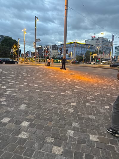 The image depicts a street scene in Basel, Switzerland, at dusk. The foreground is dominated by a cobblestone pavement, with individual stones varying in color from dark gray to lighter shades. In the midground, a street intersects with pedestrian walkways. Several streetlights with warm yellow illumination are visible, casting pools of light on the pavement. A dark-colored station wagon is parked on the left side of the street. Further down the street, traffic lights are illuminated, with some showing red signals. Two people are standing on a pedestrian crossing, one on a scooter and the other beside them. Two other individuals are further down the street, also near a pedestrian crossing. In the background, buildings of varying architectural styles line the street. One prominent building features a blue facade with white details, and another is a modern white building with a red sign on top. The sky is overcast with dark clouds, typical of twilight conditions. The overall impression is of a bustling urban environment transitioning from day to night.