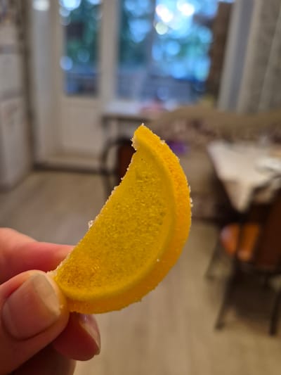 A close-up shot shows a person holding a crescent-shaped, candied orange peel slice between their fingers. The peel is a vibrant yellow-orange color and is dusted with sugar crystals, giving it a slightly textured appearance. The surface of the peel is translucent, and some small air bubbles are visible within its sugary matrix. The hand holding the peel has manicured fingernails, and the skin appears to be of light to medium tone.
The background is out of focus, suggesting a shallow depth of field. To the left, a blurred window reveals a bright, outdoor scene with dappled light and greenery, indicative of daytime. The lighting suggests indoor ambient light mixed with daylight coming through the window. The overall mood appears casual and relaxed, with no specific event or emotion being conveyed. The setting appears to be domestic, possibly a kitchen or living area, given the blurred elements in the background that could be furniture or household items. No text is visible in the image. The location is specified as Nyagan, Russia.