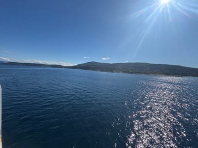 The image depicts a bright, sunny day over a body of water, likely a fjord or strait, in Hierenjárgga - Korsnes, Norway. The sky is a clear, deep blue, with the sun positioned high in the upper right corner, casting bright rays and reflections on the water's surface. The water is a rich, dark blue, with gentle ripples creating shimmering highlights from the sunlight.
In the distance, a chain of low, green hills and mountains forms the coastline. These hills appear forested and rugged, with some rocky outcrops visible. Scattered along the shoreline, especially towards the right side of the frame, are signs of human habitation, likely small houses or buildings nestled amongst the trees. The overall impression is one of a serene, natural landscape with a touch of coastal settlement. The viewpoint appears to be from a moving vessel, as indicated by the slight tilt of the frame and the presence of a white railing along the left edge of the image, suggesting the photo was taken from a boat or ferry. There are no people visible in the scene.
