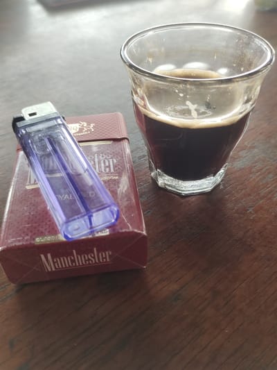 This is a close-up, overhead shot of a cigarette pack and a glass of coffee on a wooden table. The cigarette pack is maroon with gold lettering that reads "Manchester" and "ROYAL RED". A clear plastic lighter with a purple tint rests on top of the cigarette pack. The glass contains dark coffee with a layer of crema on top. The lighting suggests an indoor setting, and the wooden table has a visible grain. There is no indication of the specific location within Mee, Indonesia.